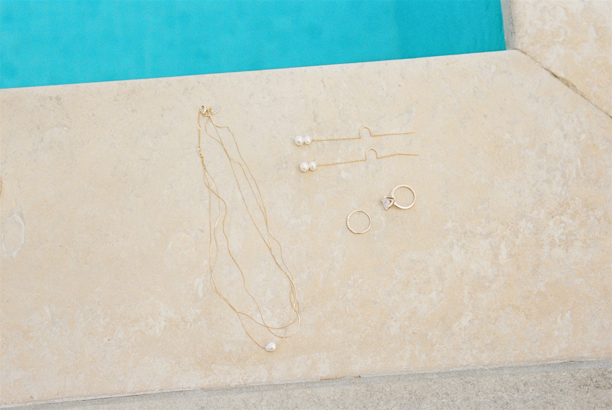 A bride's jewelry lays on the edge of a pool with gold and pearls