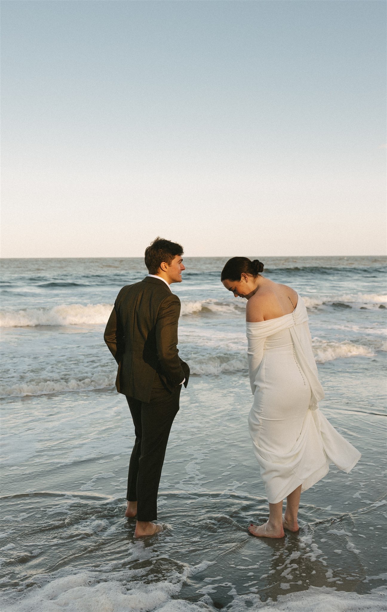 A bride and groom walk barefoot in the low tide waters of one of the Charleston wedding venues beaches at sunset