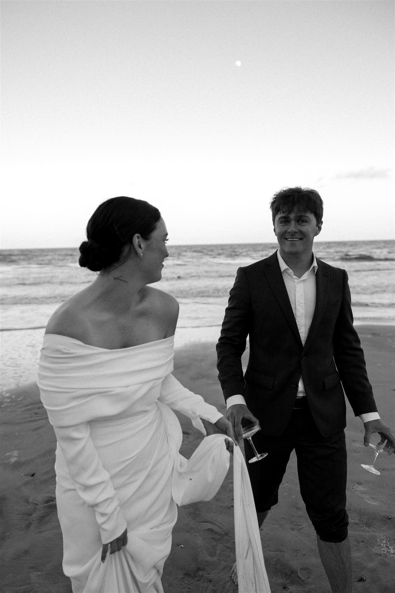 Newlyweds smile while walking together on a beach as groom carries drink glasses in black and white at one of the Charleston wedding venues