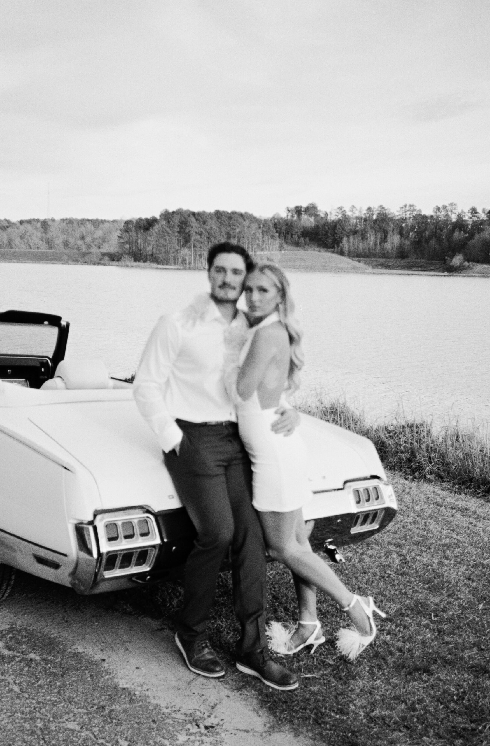 Newlyweds lean on the trunk of a vintage muscle car parked by a lake