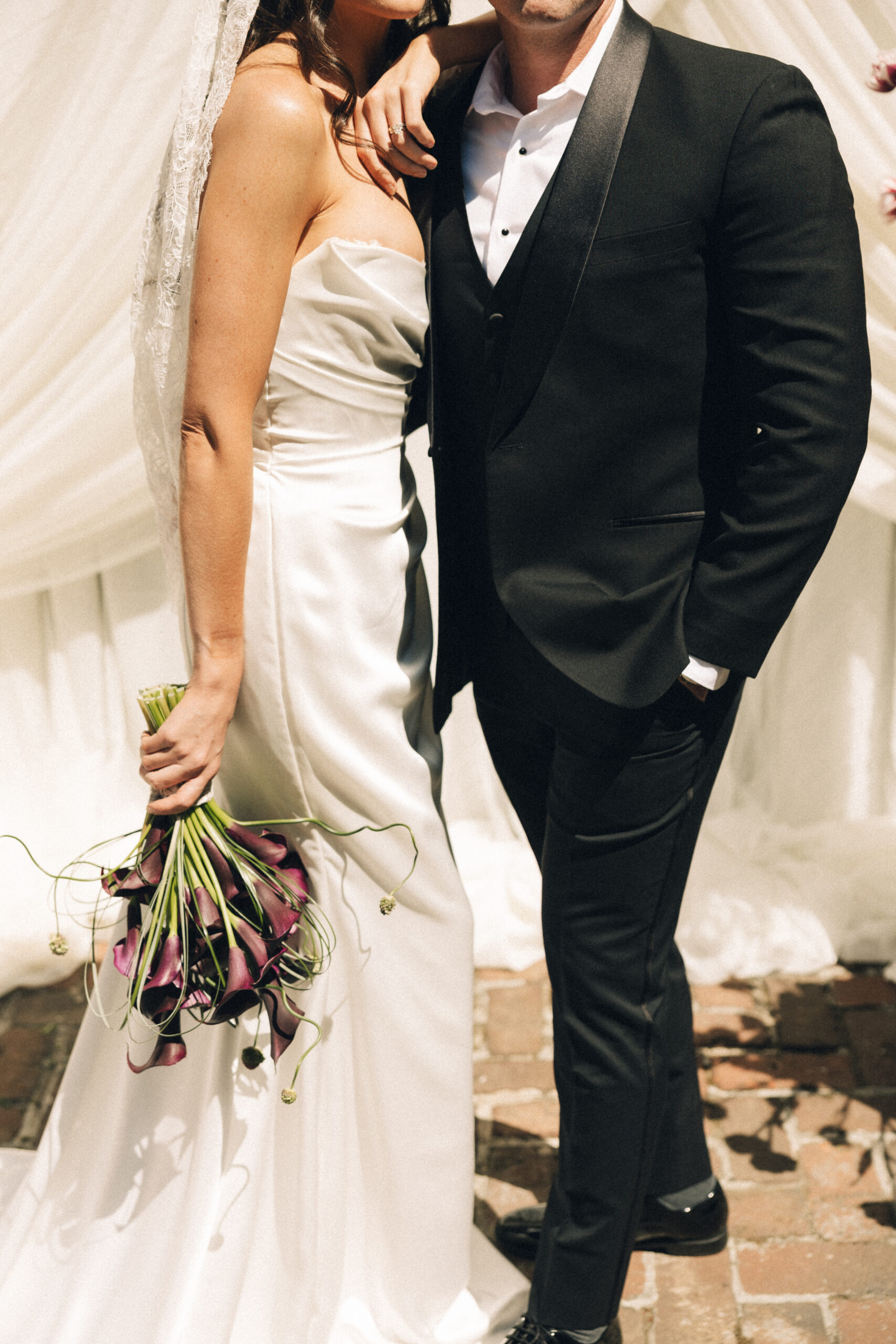 A bride leans on the shoulder of her groom in a black suit with hand in pocket outdoors on a brick patio