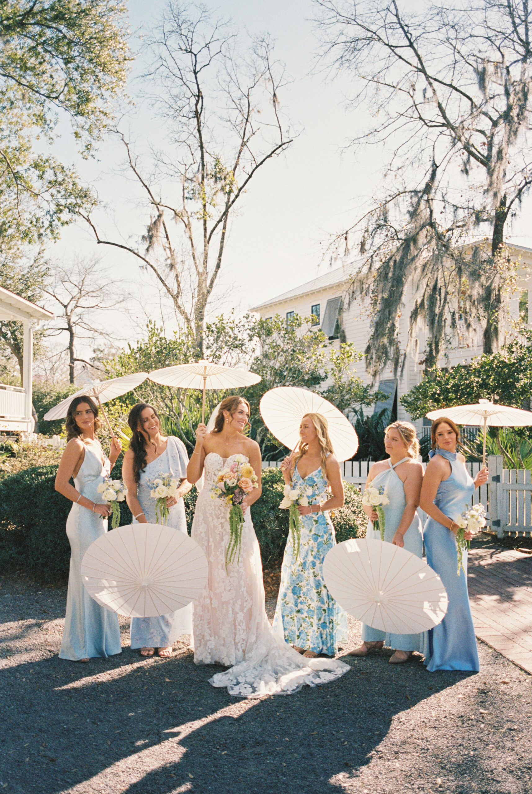 A bride laughs with her bridesmaids in blue dresses and holding parasols in the garden of one of the Charleston wedding venues