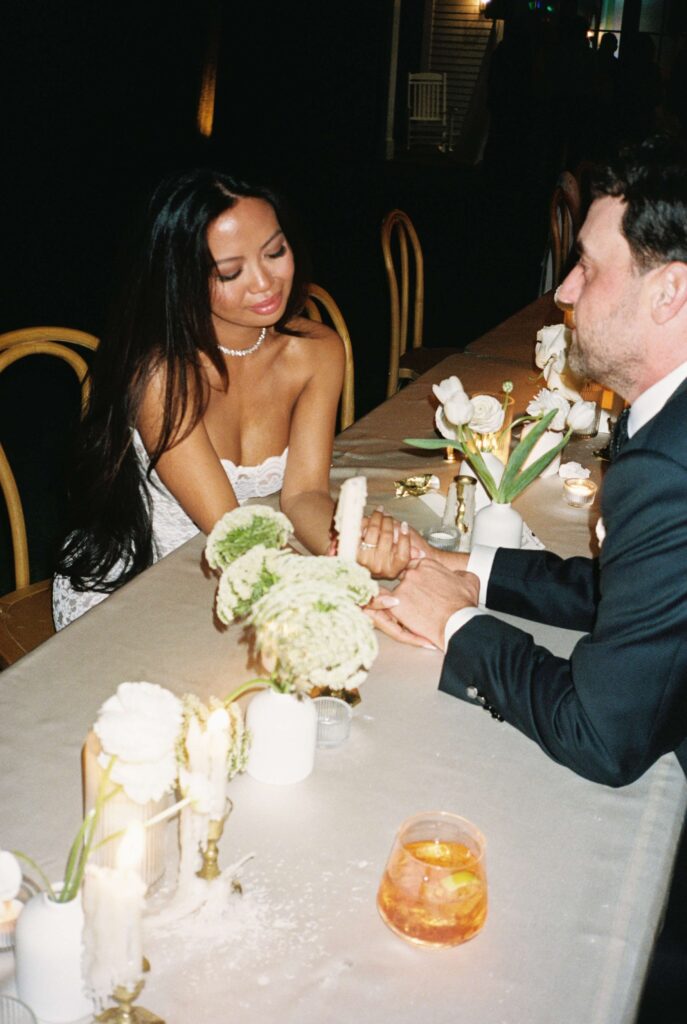 Newlyweds hold hands over a reception table under candle light