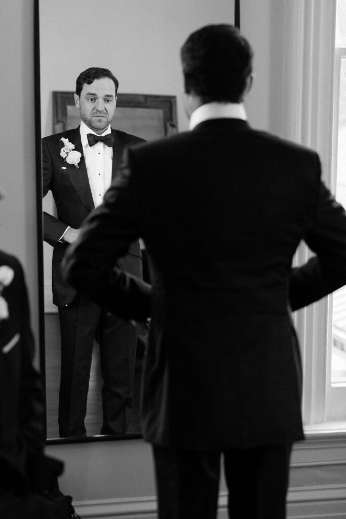 A groom adjusts his jacket while standing in a mirror