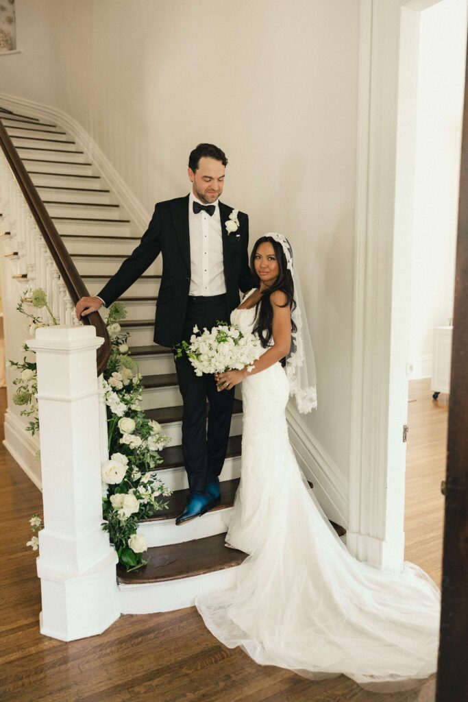 newlyweds stand on the grand staircase adorned with white flowers at their merrimon wynne house wedding