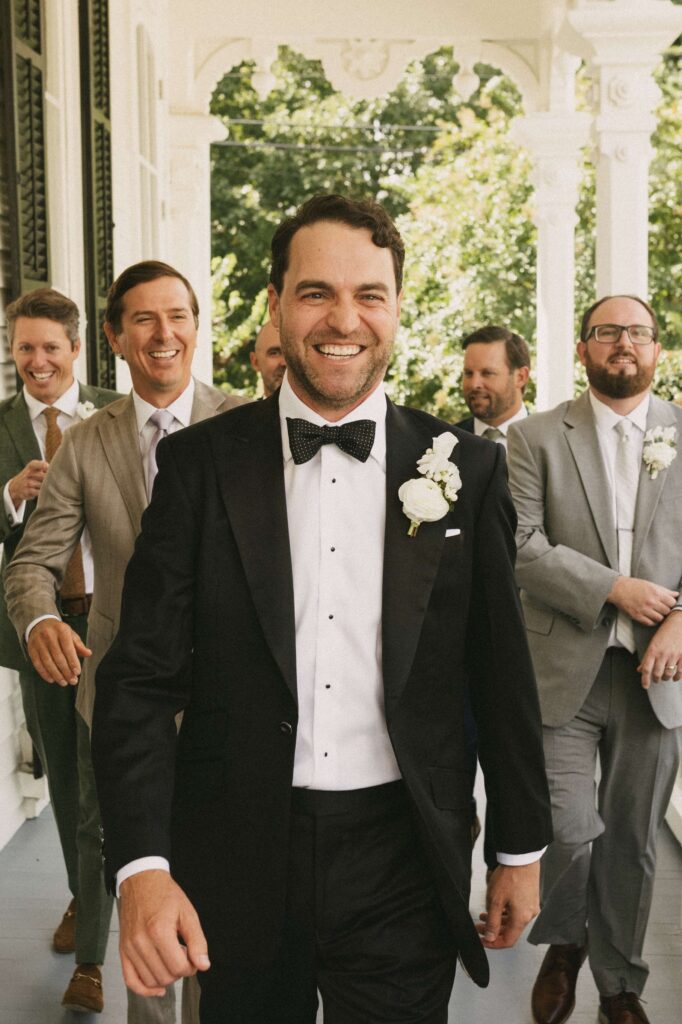 A laughing groom walks on the merrimon wynne house wedding venue porch with his happy groomsmen