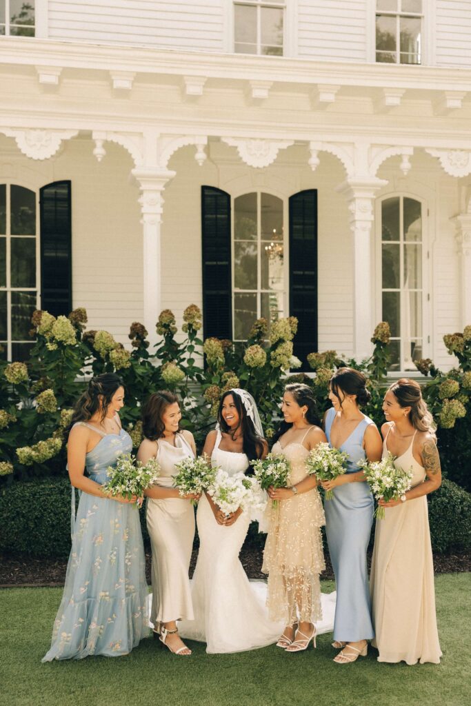 A bride laughs with her bridesmaids all holding white bouquets out front of the merrimon wynne house wedding