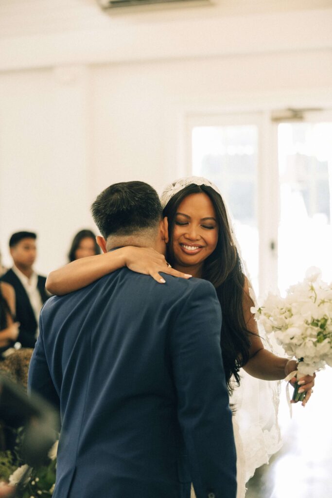 A bride smiles while hugging a man at the altar in a blue suit