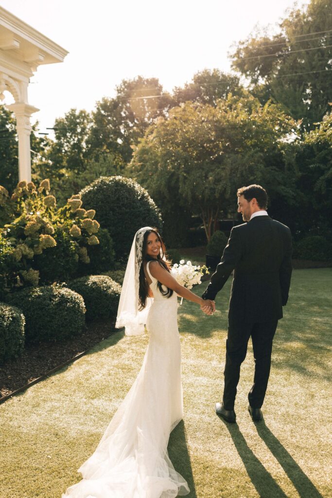 A happy bride smiles back over her shoulder while walking the merrimon wynne house wedding venue lawn holding hands with her groom