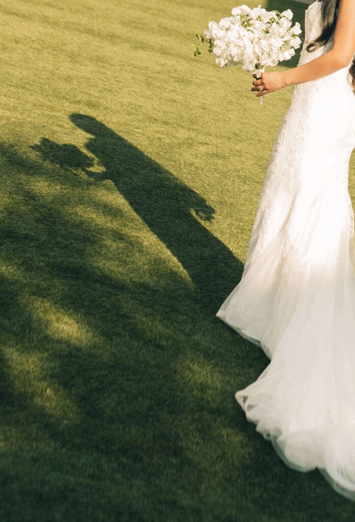 Details of a bride's shadow as she walks in the manicured lawn holding bouquet