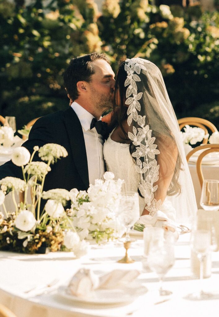 A bride and groom kiss while sitting at the outdoor reception table