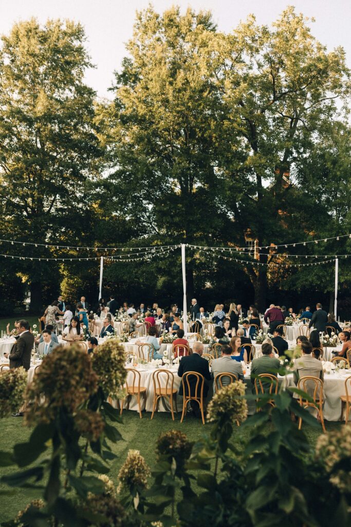 A peek through the flowers of a busy merrimon wynne house wedding receptions outdoors in the garden under market lights