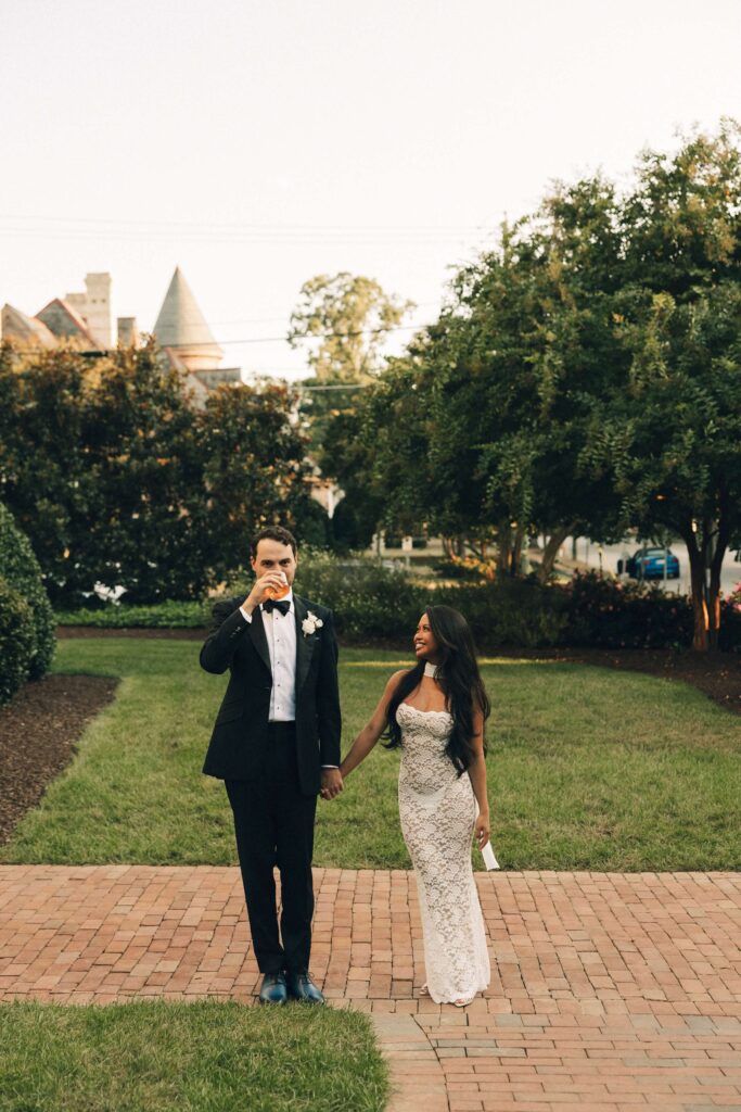A groom sips a drink while holding hands with his happy bride at their merrimon wynne house wedding