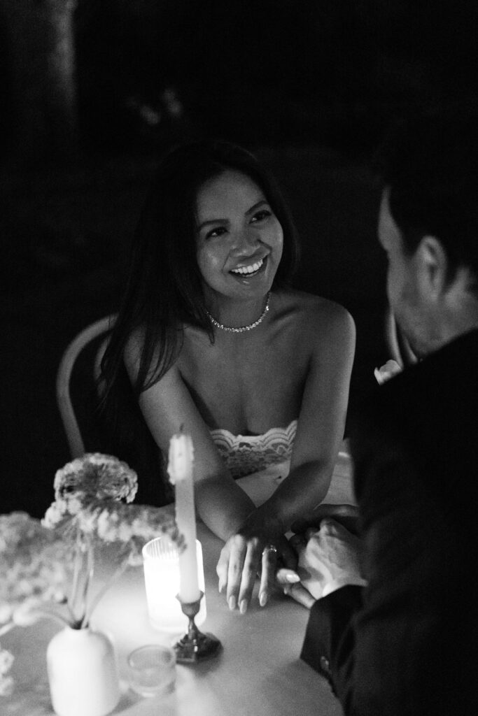 A bride smiles while sharing a quiet moment in black and white at a merrimon wynne house wedding reception table holding hands with her groom