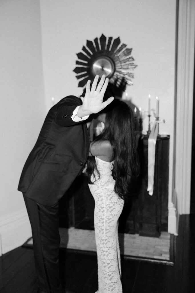 A groom puts his hand out while kissing his bride in a hallway in black and white