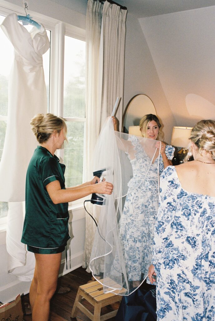 Bridesmaids help steam the veil as the dress hangs in a window
