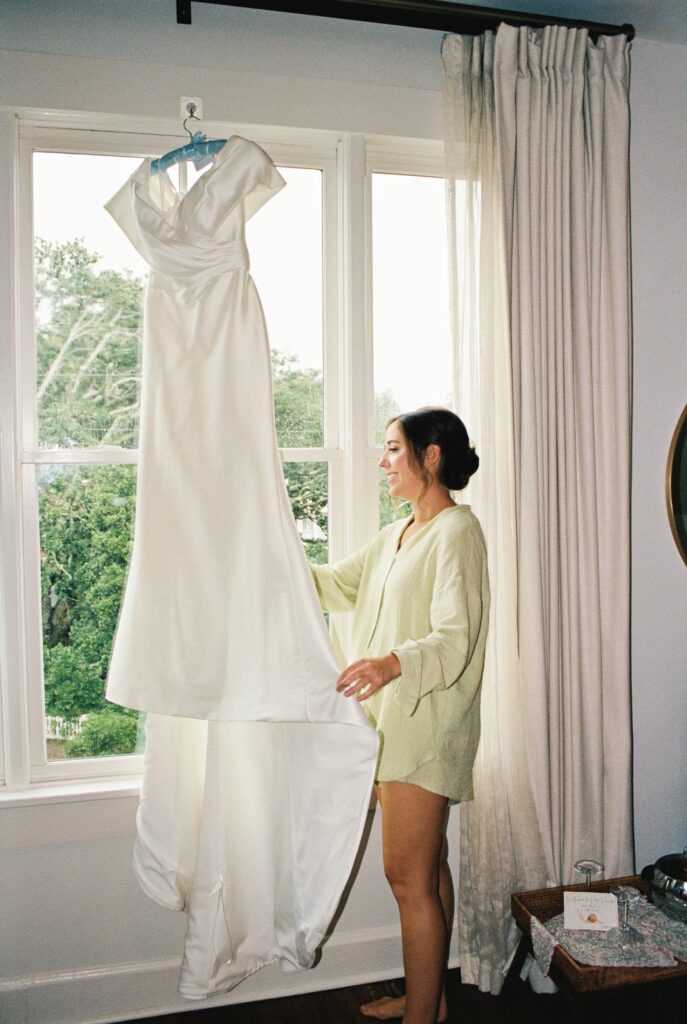 A happy bride in green pajamas smiles while admiring her gown hanging in a window at her Post House Inn wedding