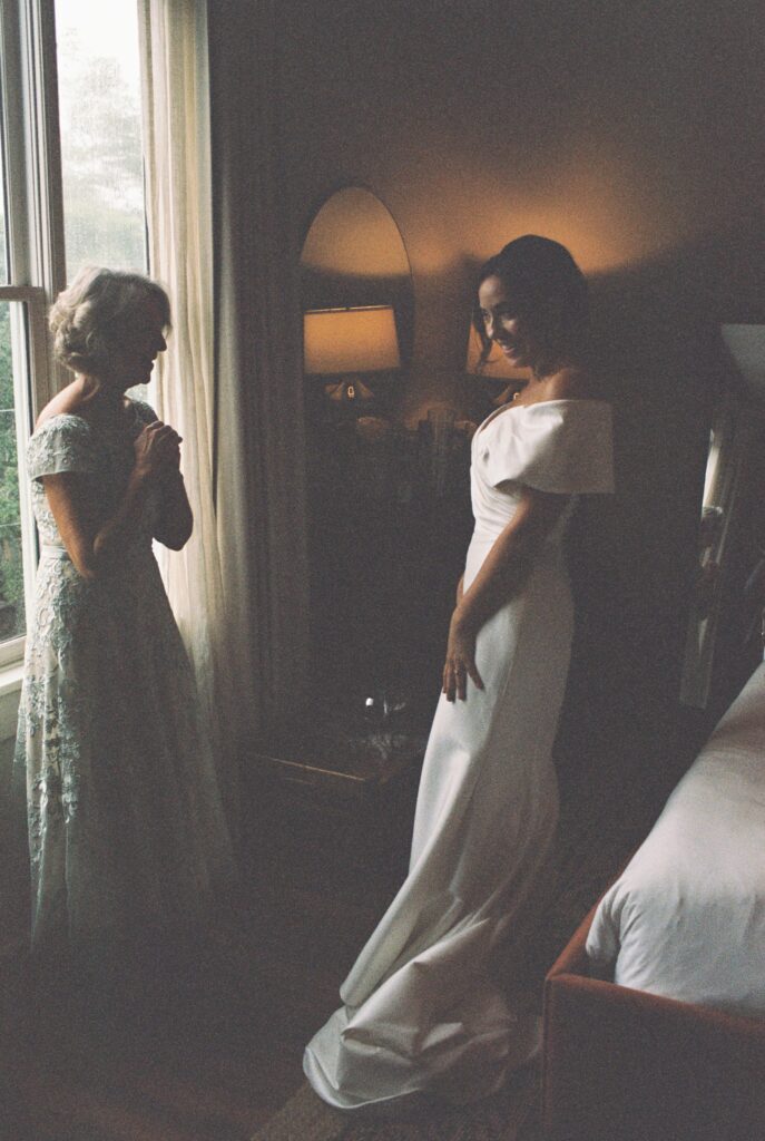 A smiling bride shows off her gown to mom while standing in a window of the Post House Inn wedding venue