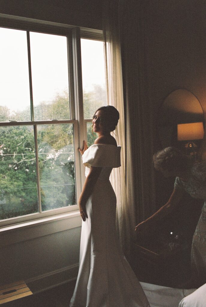 A happy bride smiles over her shoulder while standing in a window in her silk gown at the Post House Inn wedding venue getting ready suite