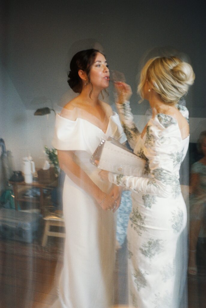 A bridesmaid in a floral print dress helps the bride with her lip makeup in the getting ready room
