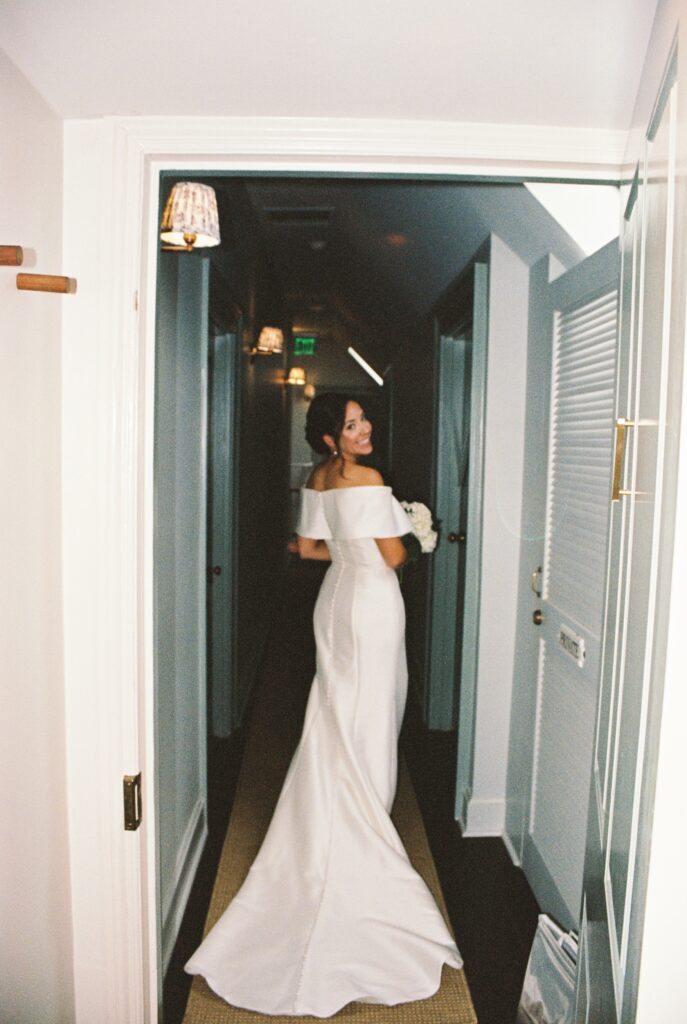 A happy bride holds her bouquet and smiles big over her shoulder while heading down a hallway to her Post House Inn wedding ceremony