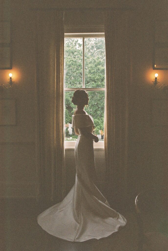 A bride stands in silhouette in a window wearing her glamorous silk gown at her Post House Inn wedding