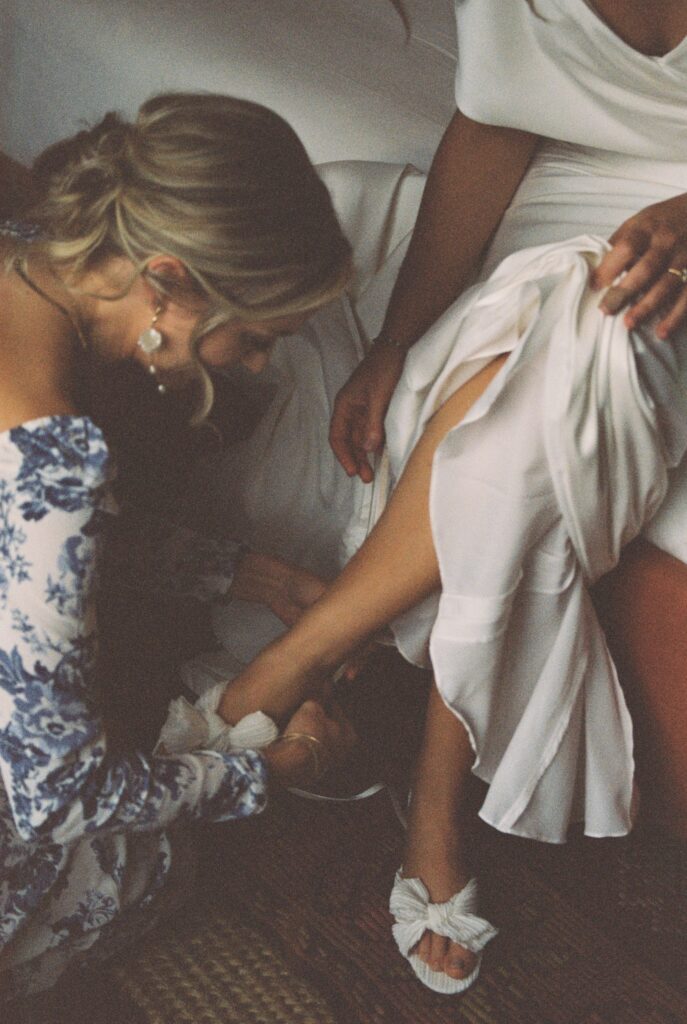 A bridesmaid in a floral print dress helps the bride put on her shoes
