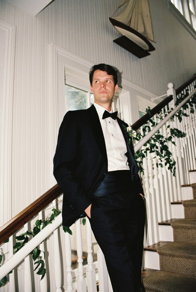 A groom leans on the stairs railing with hands in pockets in a black tuxedo during his Post House Inn wedding
