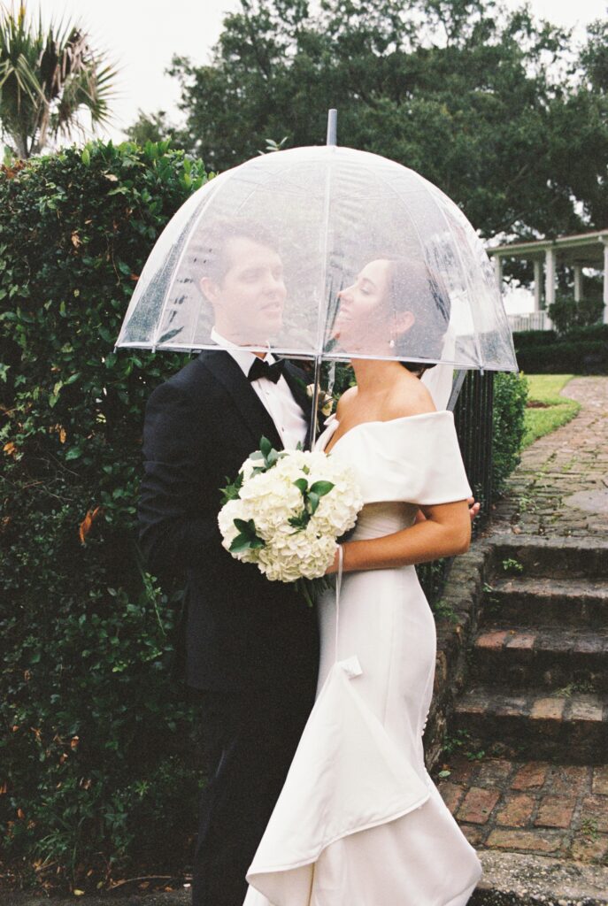 Newlyweds laugh and snuggle under a clear umbrella while exploring the Post House Inn wedding venue grounds