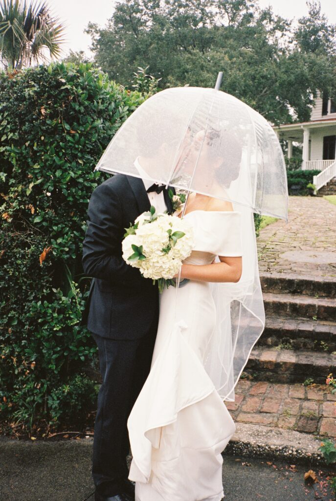 A bride and groom kiss under a clear umbrella while holding the white bouquet in the gardens