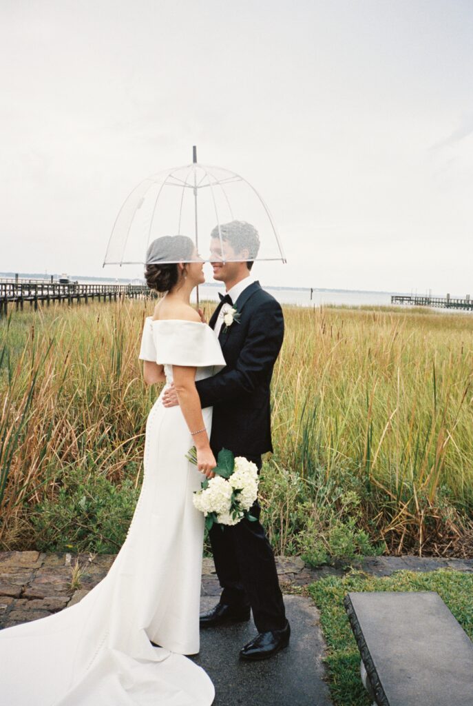 Newlyweds smile at each other while hugging under a clear umbrella in the rain by the water during their Post House Inn wedding