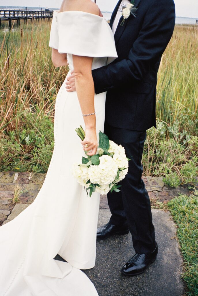 Details of a bride holding her white bouquet by her side while cuddling her groom on a sidewalk by the water