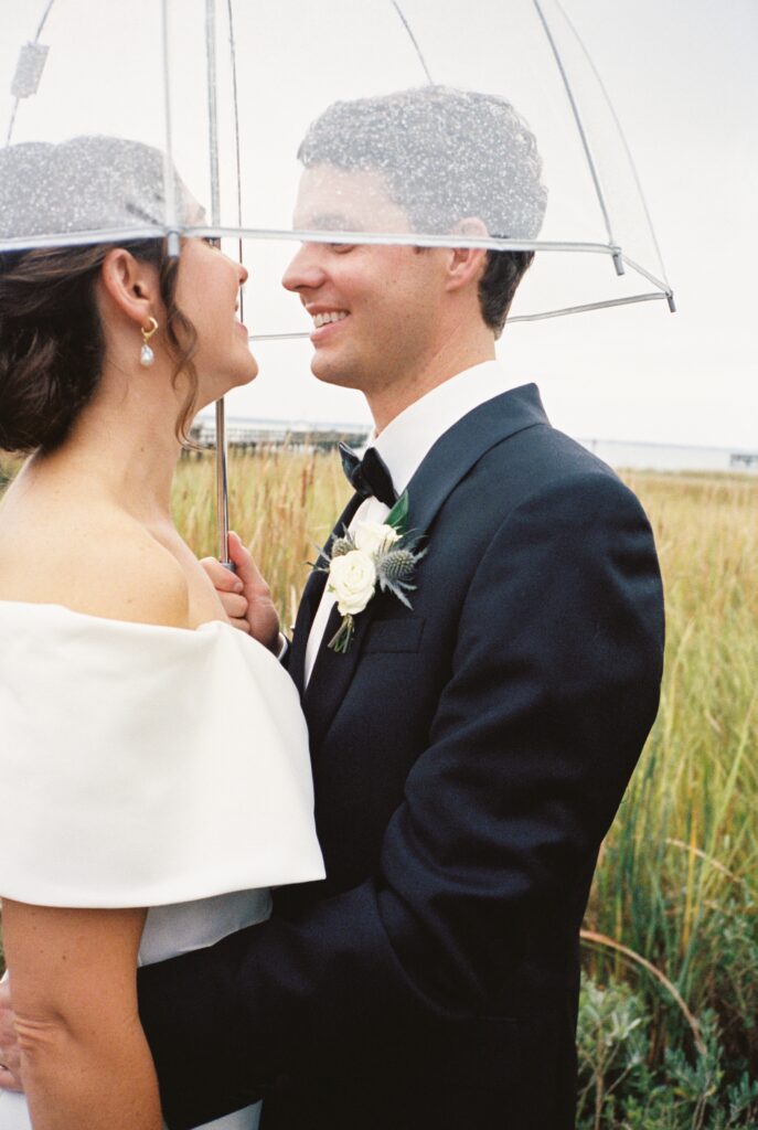 A bride and groom smile and lean in for a kiss under a clear umbrella in a field of tall grass at their Post House Inn wedding