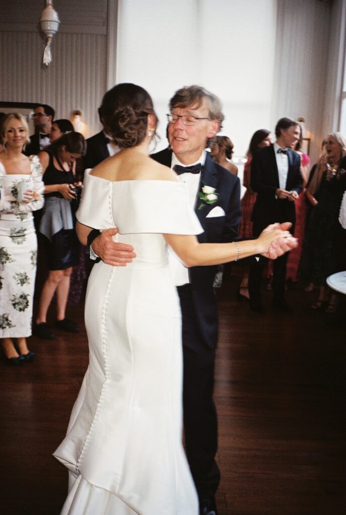A bride dances with dad during father daughter dance at her reception as guests smile looking on