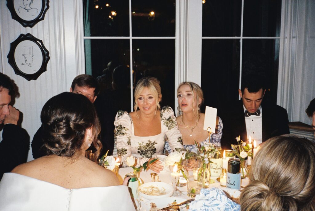 Two bridesmaids listen at the dinner table to the bride talking to them covered in candles and white roses