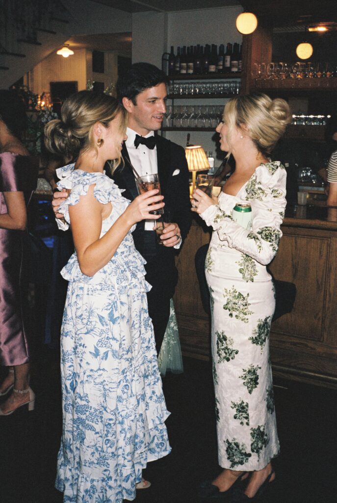 A man in a black tuxedo chats by the bar with two women in floral print dresses at a wedding