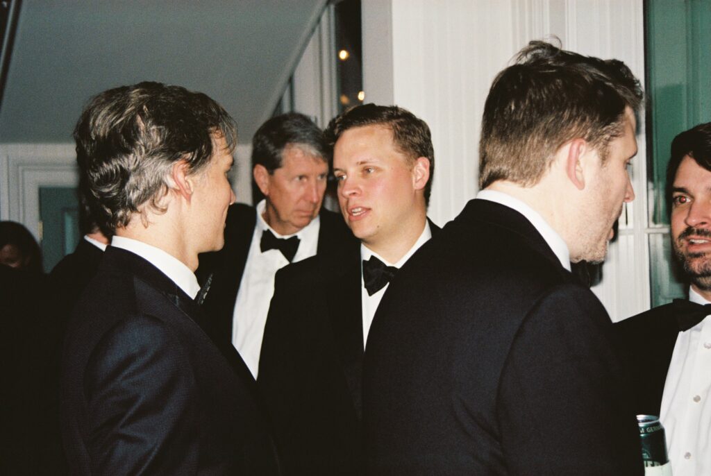A group of men in black tuxedos chat and laugh over drinks at a wedding reception