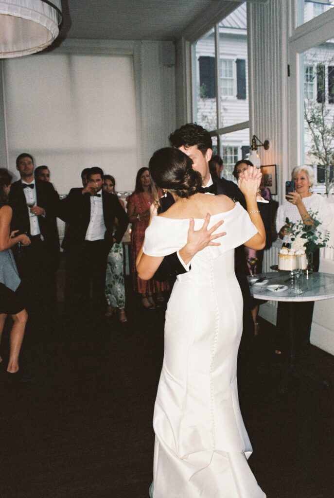 A bride and groom dance close as guests watch and film at their indoor reception