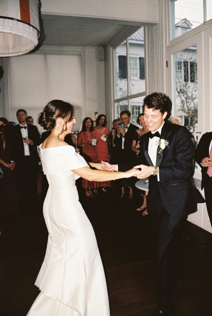 Newlyweds share a laugh while holding hands and dancing surrounded by guests at their reception