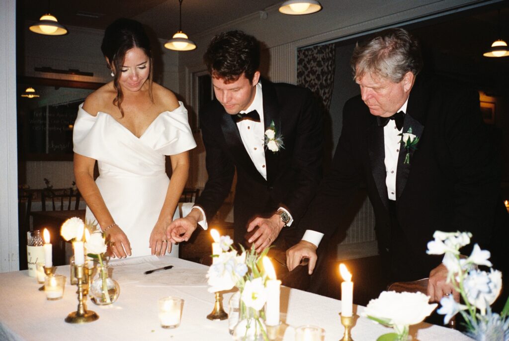 A bride and groom prepare to sign their marriage license on a white table with candles and flowers