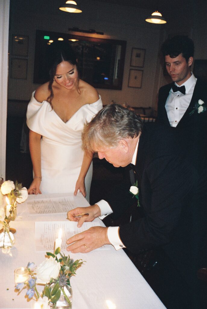 A man leans over to sign a marriage license as the bride and groom look on