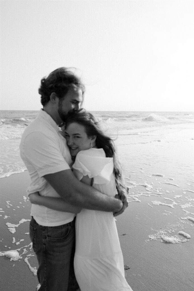 Newlyweds snuggle on a windy beach in black and white by the water