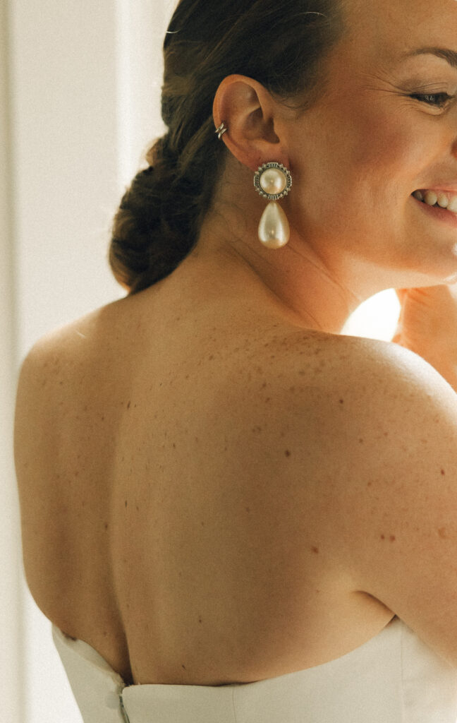 Details of a bride's large pearl earring as she smiles putting on the other
