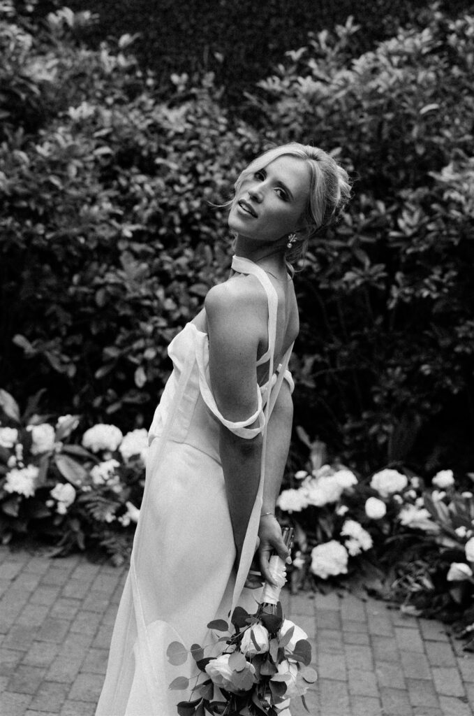 A bride holds her bouquet behind her back while smiling over her shoulder in a garden at one of the Wilmington wedding venues