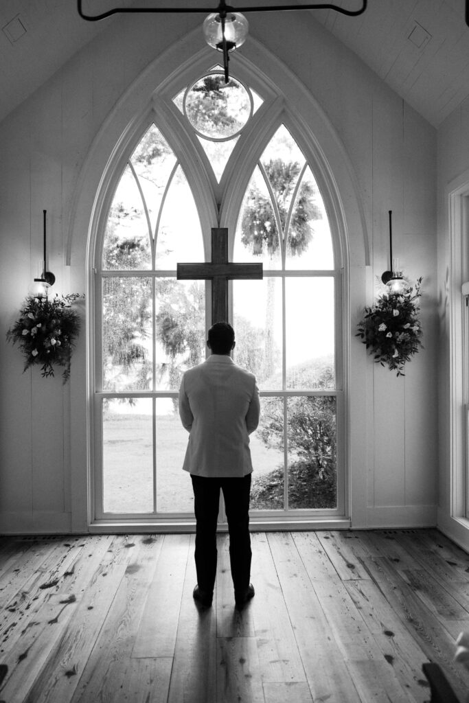 A groom stands looking up to the cross in a white tux jacket in a chapel