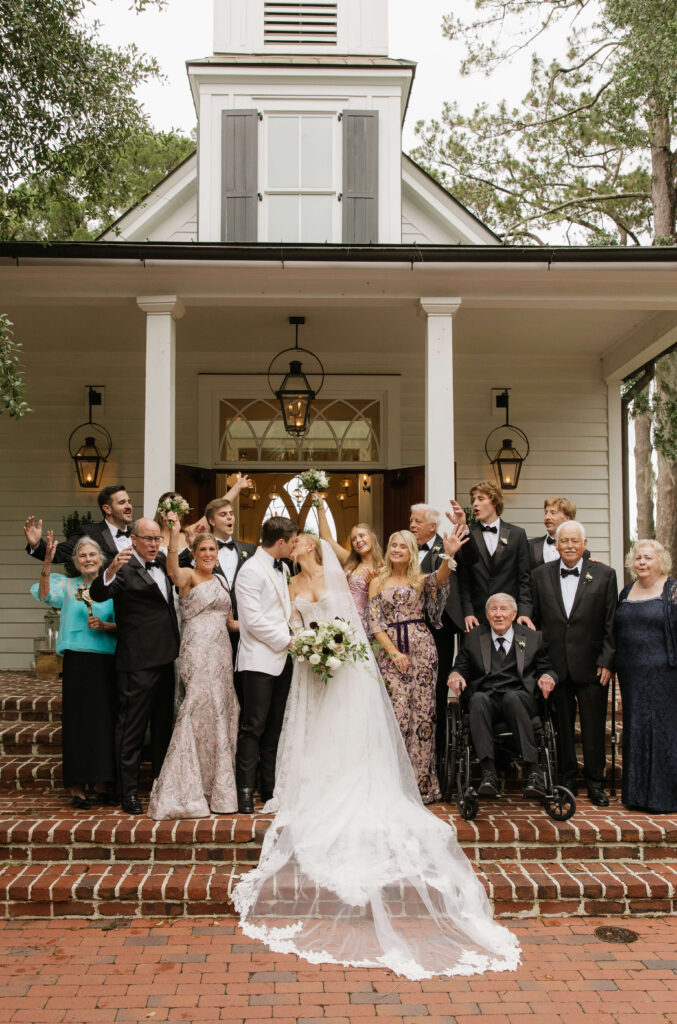 Newlyweds kiss on the front porch steps with family all celebrating around them at their montage palmetto bluff wedding