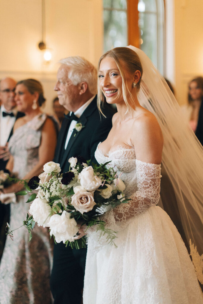 A bride smiles while walking down the aisle with dad and a large white bouquet
