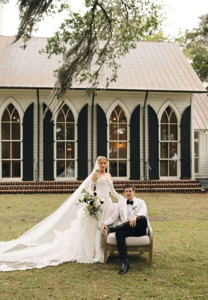 A groom sits in a chair in his white tux jacket in a lawn as his bride stands next to him with a long flowing veil and train at their montage palmetto bluff wedding