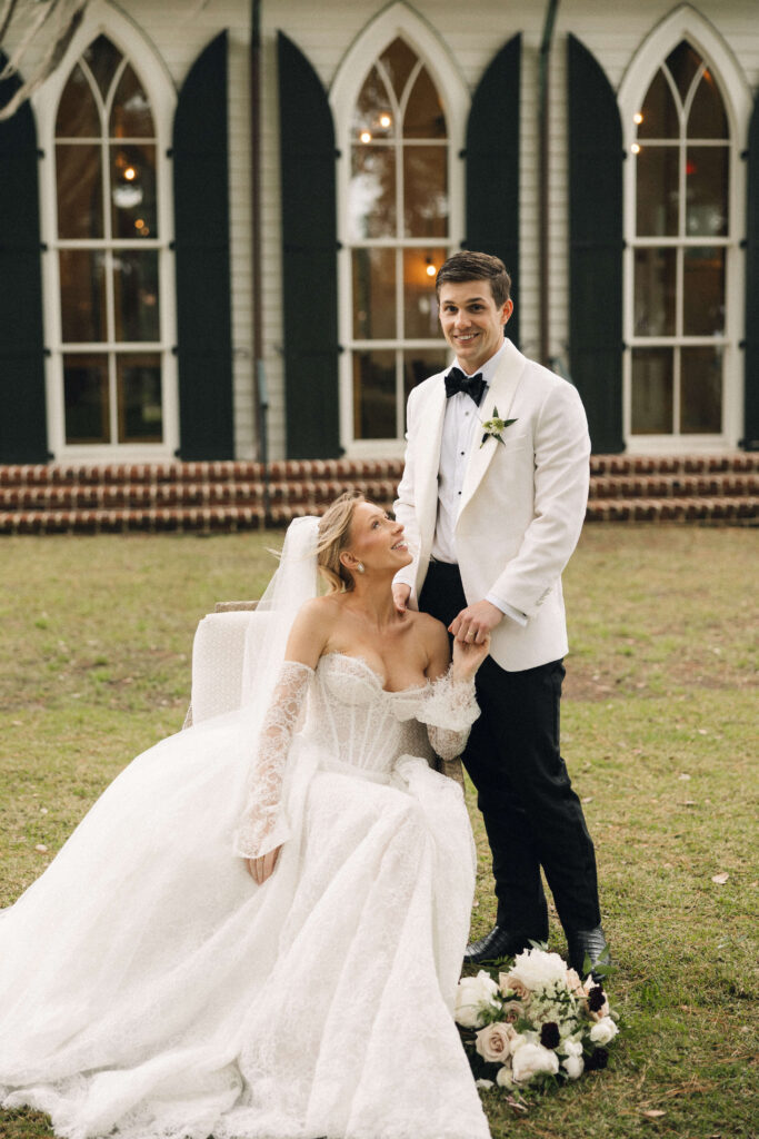 A bride sits in a chair in a lawn while smiling up to her groom in a white tux jacket outside the montage palmetto bluff wedding venue