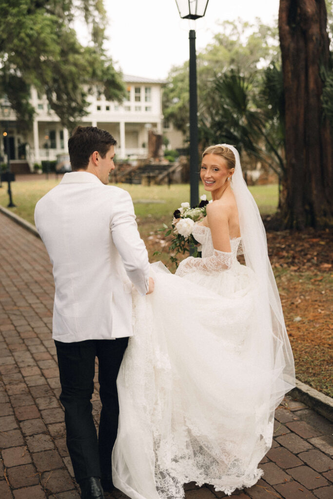 A bride smiles over her shoulder while walking on a sidewalk as her groom holds her train at their montage palmetto bluff wedding